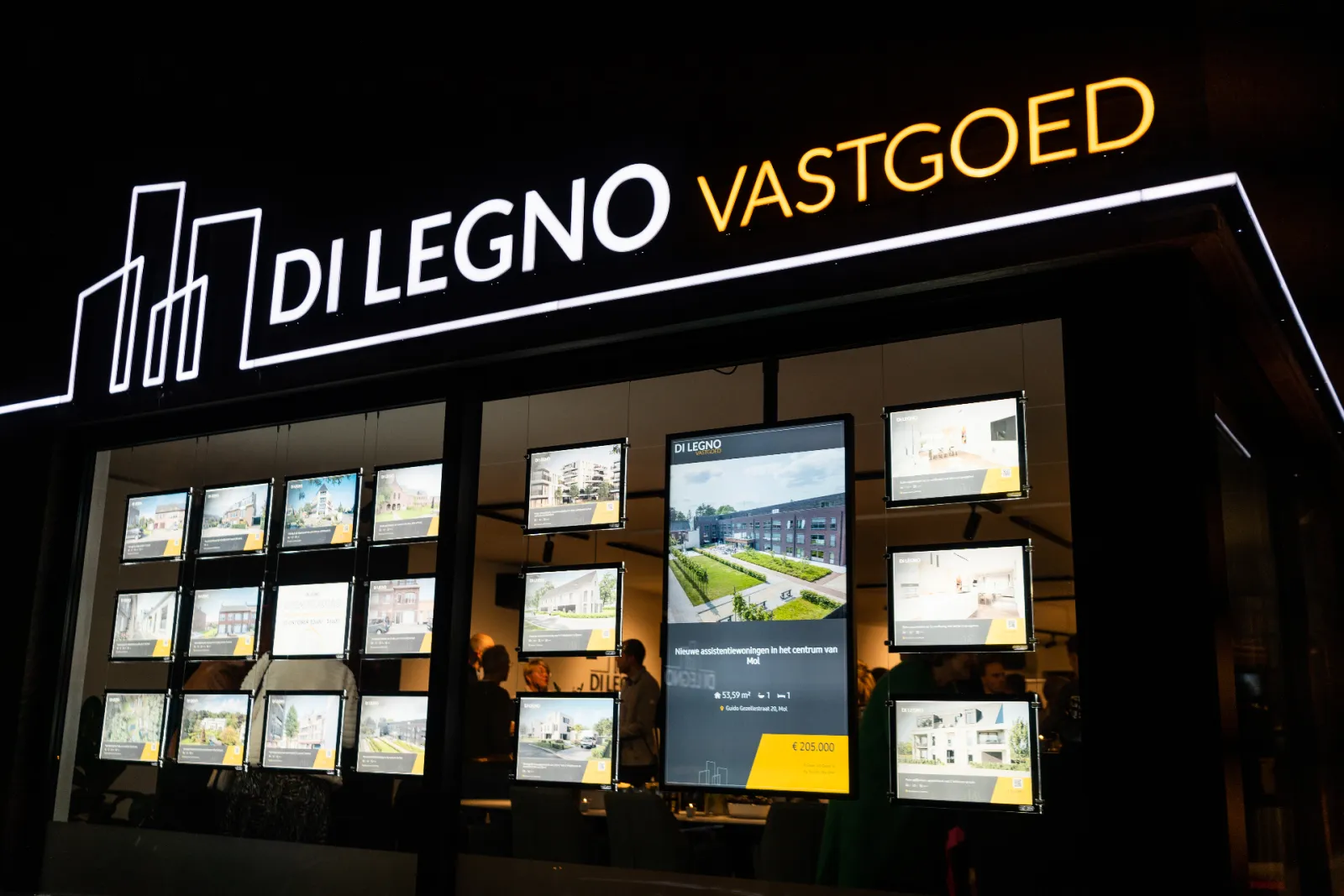 The lit shop window of Di Legno Vastgoed in the evening, with a portrait screen among the properties