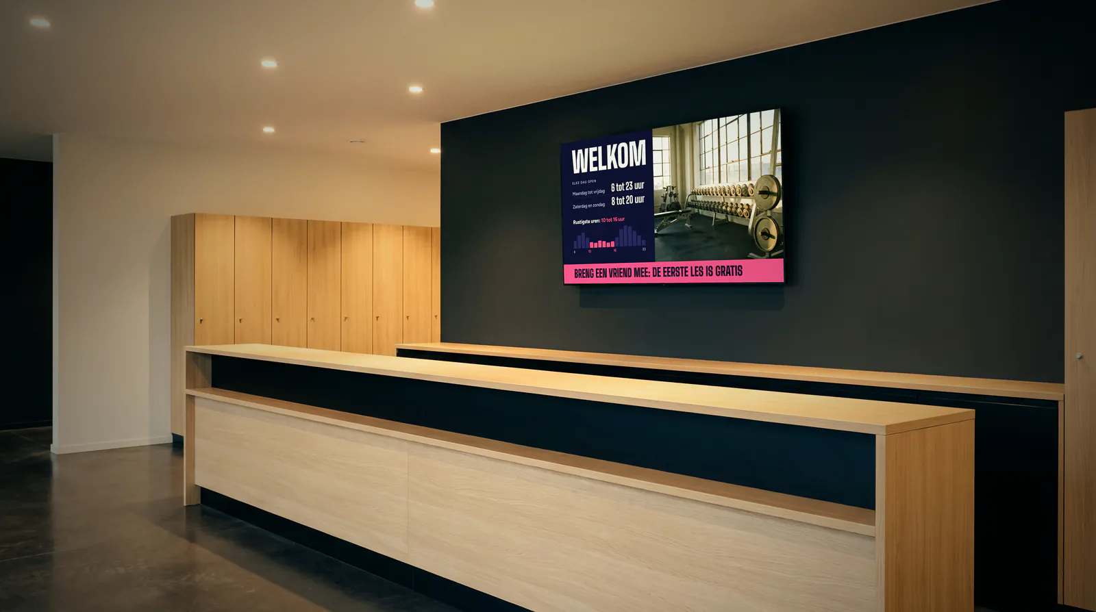 Onthaal van een sportschool met een lange balie in licht hout, houten lockers en een donkere muur met een scherm dat welkom heet en de openingsuren toont