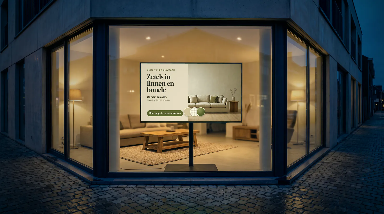 Immeuble d'angle d'un magasin de meubles à la tombée du jour, avec dans la vitrine un écran sur pied devant un salon avec un canapé et une table basse en bois